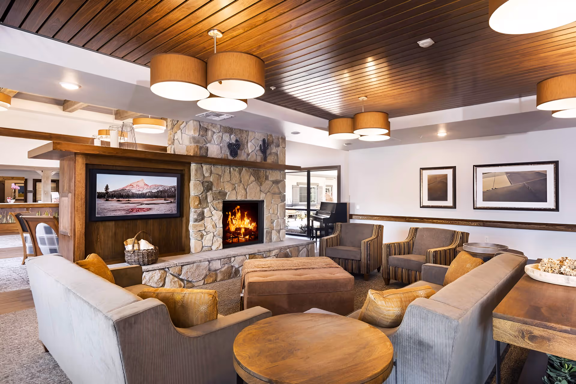 Cozy communal living room with a stone fireplace, wall-mounted TV, sofas and armchairs under a wooden ceiling with pendant lights.