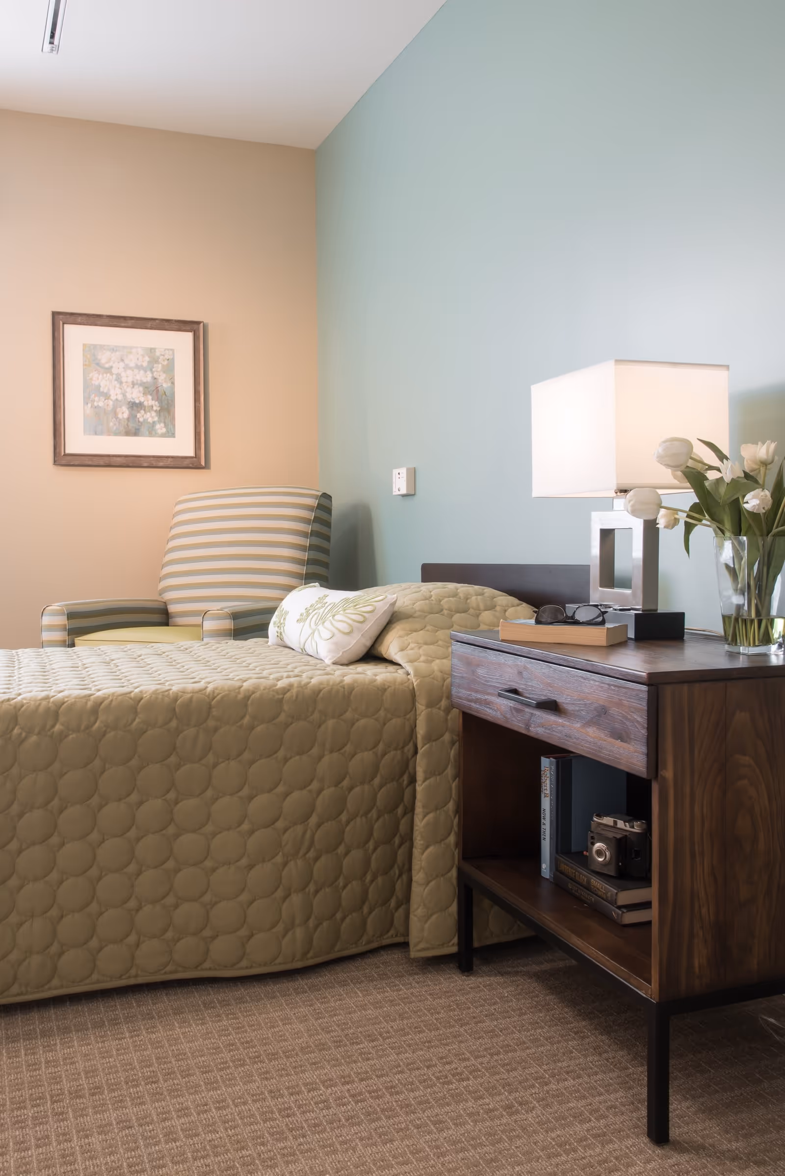 A cozy bedroom featuring a single bed with a quilted olive-green bedspread and a decorative pillow. Next to the bed is a wooden nightstand with a drawer and an open shelf holding books and a vintage camera. On top of the nightstand is a modern square-shaped lamp, a pair of glasses, a book, and a vase with white tulips. A striped armchair is positioned against the beige wall, which also has a framed floral artwork. The adjacent wall is painted light blue, and the floor is carpeted in a neutral tone.