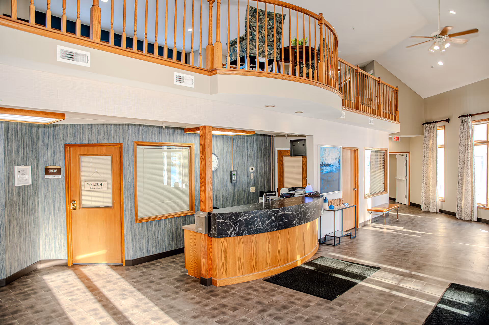 A bright and spacious reception area with a curved wooden front desk topped with black marble. Behind the desk is a wall with a door and window, and above is a wooden railing balcony. The floor is tiled, and large windows with curtains allow natural light to fill the room. There is a ceiling fan with lights and a small table with decorative items near the entrance.