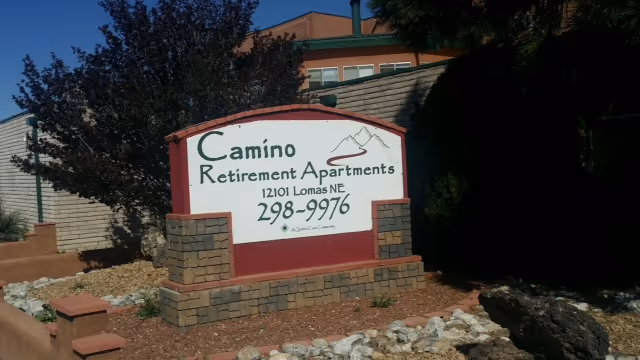 Outdoor sign for Camino Retirement Apartments located at 12101 Lomas NE with phone number 298-9976, surrounded by landscaping with rocks, bushes, and trees.