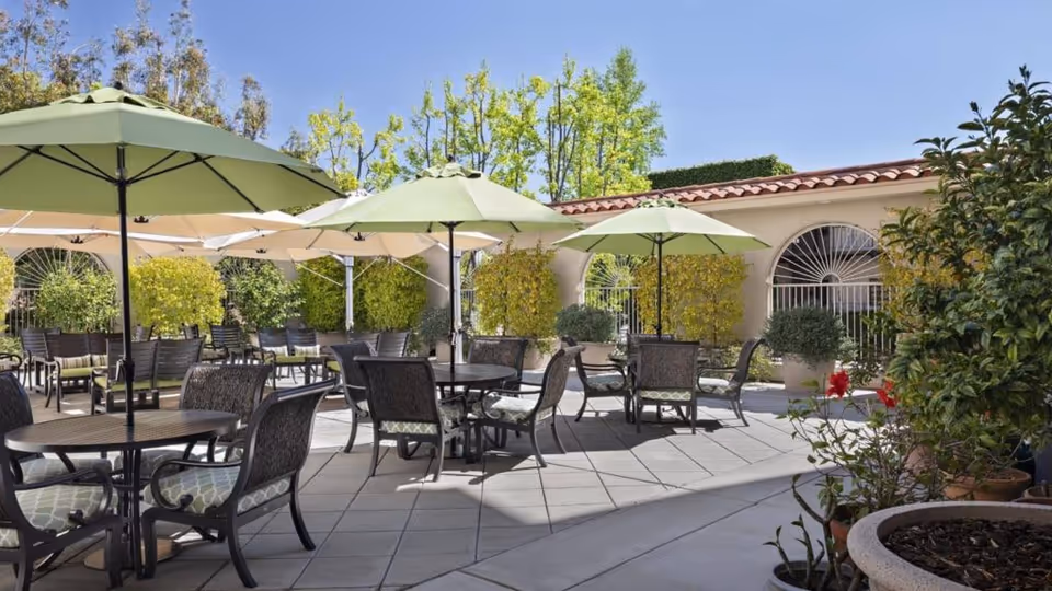Outdoor patio area with multiple round tables and cushioned chairs under large green umbrellas. The patio is surrounded by potted plants and greenery, with a building featuring arched windows and a tiled roof in the background under a clear blue sky.