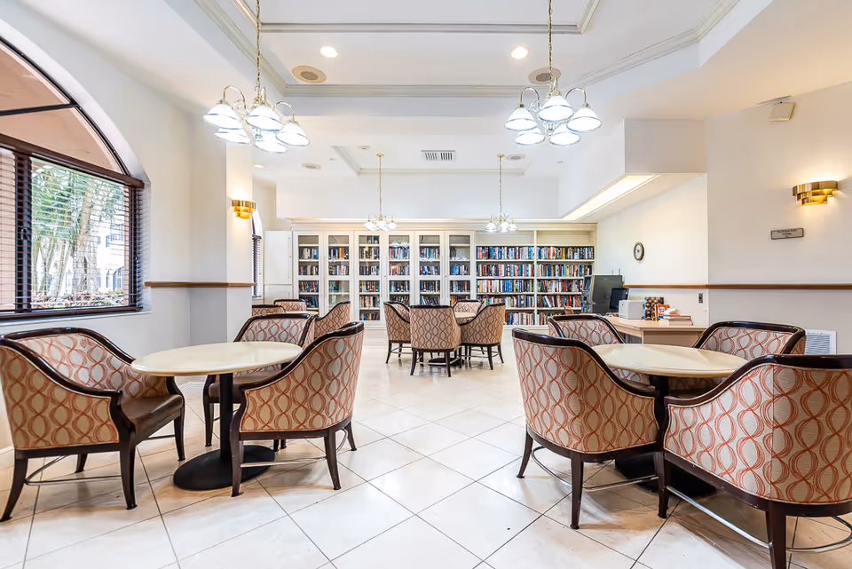 A bright and spacious senior living common area with multiple round tables surrounded by patterned upholstered chairs. The room features large windows with wooden blinds, white tiled floors, and built-in bookshelves filled with books along the back wall. Elegant ceiling lights hang from the tray ceiling, and there is a desk with a computer and some books on the right side of the room.