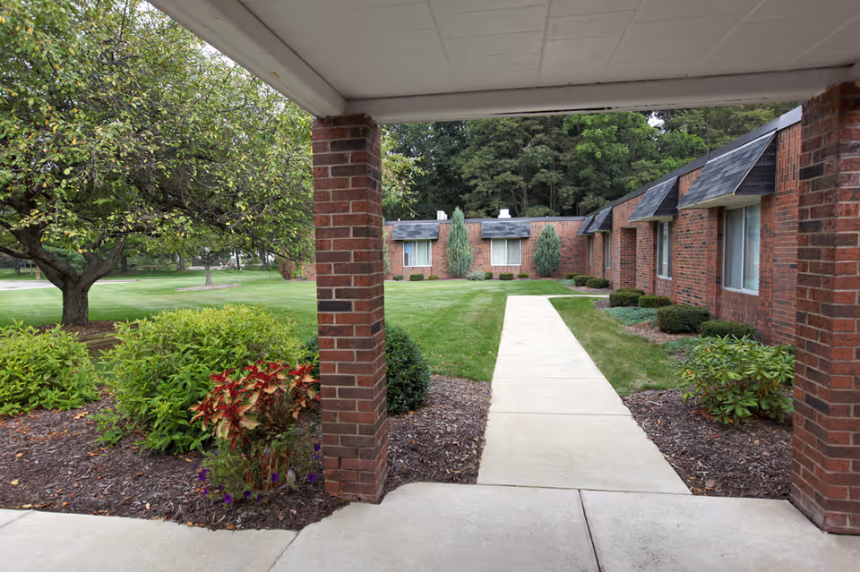 View of a landscaped courtyard with a concrete walkway leading through green grass and bushes, surrounded by a single-story brick building with windows. There is a covered patio area with brick columns in the foreground and trees in the background.