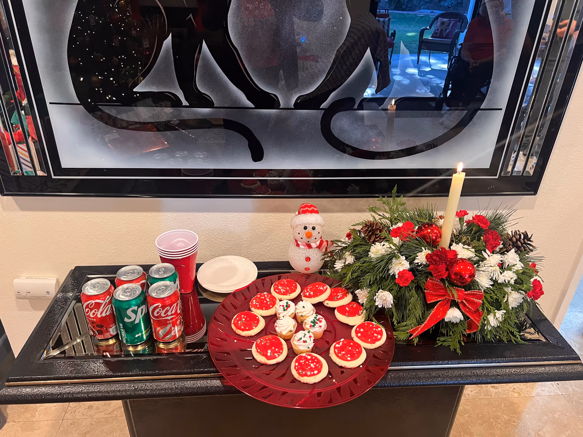 A festive holiday setup on a black table featuring a red plate with decorated sugar cookies and cupcakes, a floral arrangement with red and white flowers, pine cones, red ornaments, and a lit candle. There are also cans of Coca-Cola and Sprite, red plastic cups, and white paper plates. A decorative frosted glass panel is mounted on the wall behind the table.