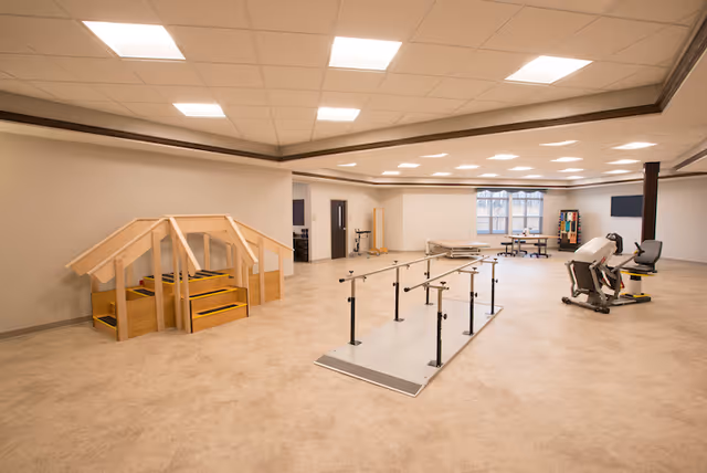 Spacious rehabilitation/therapy room with parallel walking bars, exercise machines, and wooden practice stairs under bright ceiling lights.