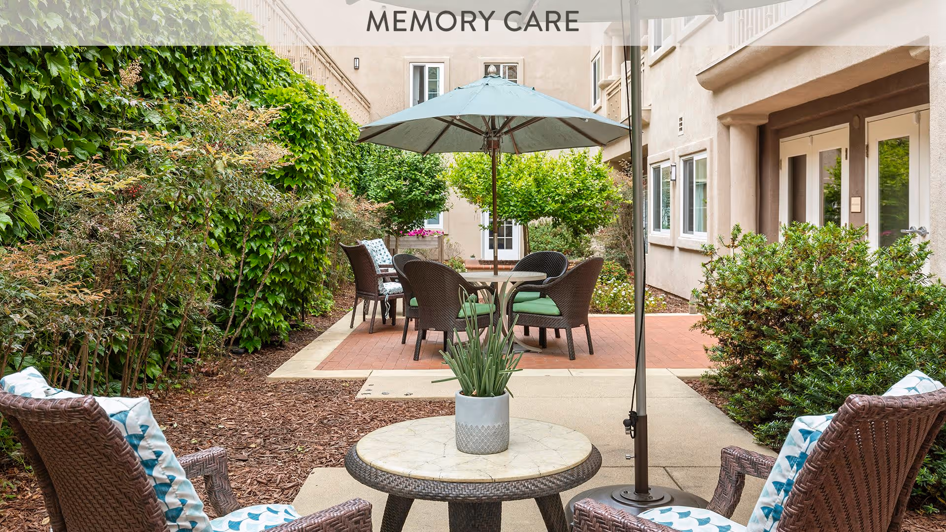 Outdoor patio area at Atria Newport Plaza with wicker chairs and a round table with a potted plant in the foreground, additional seating with cushions and an umbrella in the background, surrounded by greenery and bushes, with a building entrance visible.