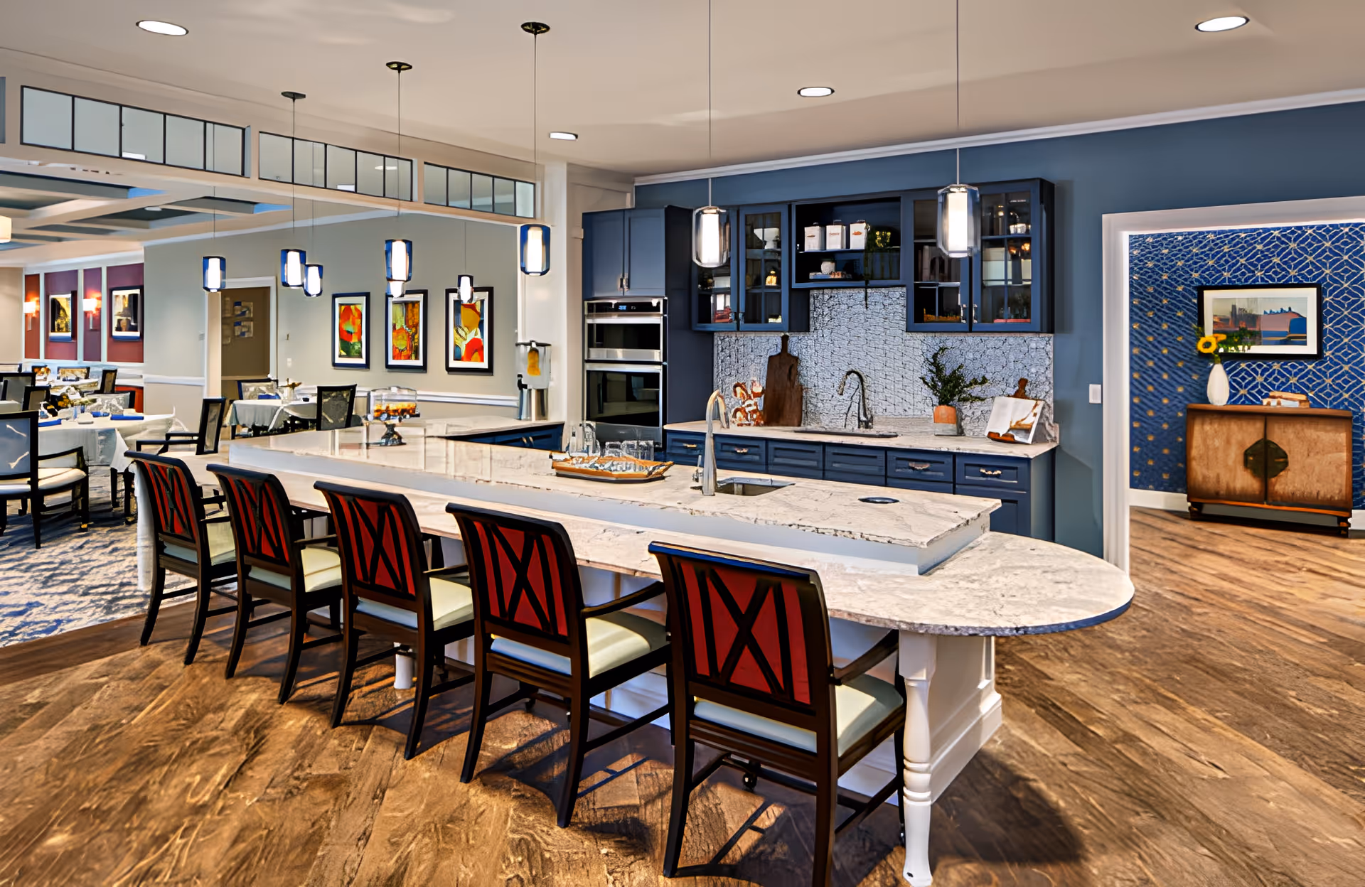 A modern kitchen and dining area in a senior living facility with a long marble countertop island featuring six chairs with red backs and light cushions. The kitchen has blue cabinets, a built-in oven, a sink, and pendant lights hanging from the ceiling. In the background, there is a dining area with tables and chairs, colorful framed artwork on the walls, and a blue accent wall with a wooden cabinet and decorative items.