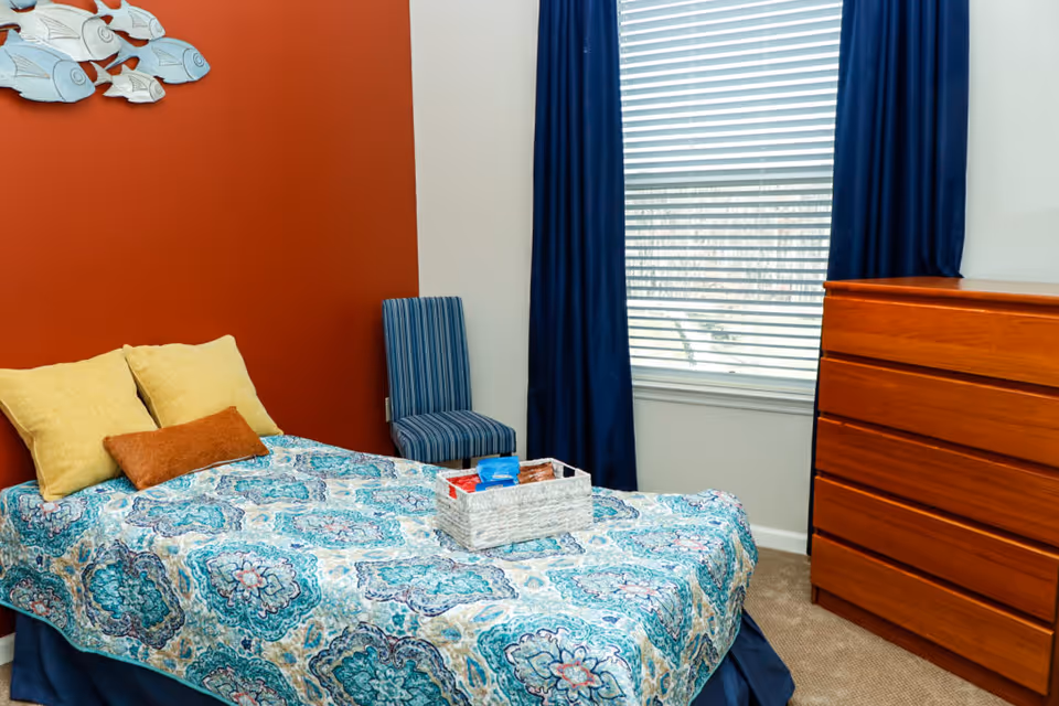 A bedroom with a bed covered in a blue and white patterned bedspread, two yellow pillows, and one brown pillow. A wicker basket with items is placed on the bed. There is a blue striped chair next to the bed, a wooden dresser with four drawers, a window with white blinds and dark blue curtains, and a red accent wall with a decorative metal fish artwork.
