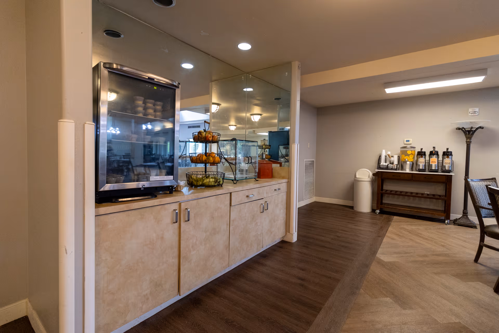 Interior communal dining/lounge area featuring a snack and beverage station with a refrigerated display, tiered fruit baskets, cabinets, and seating.
