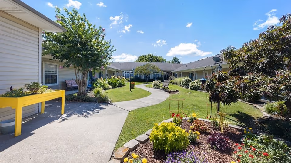 A sunny outdoor courtyard at Brookdale Goodlettsville featuring a winding concrete pathway surrounded by green grass, colorful flower beds, and various trees and shrubs. There are benches along the path and a yellow raised planter box near the building. The sky is clear with a few clouds.