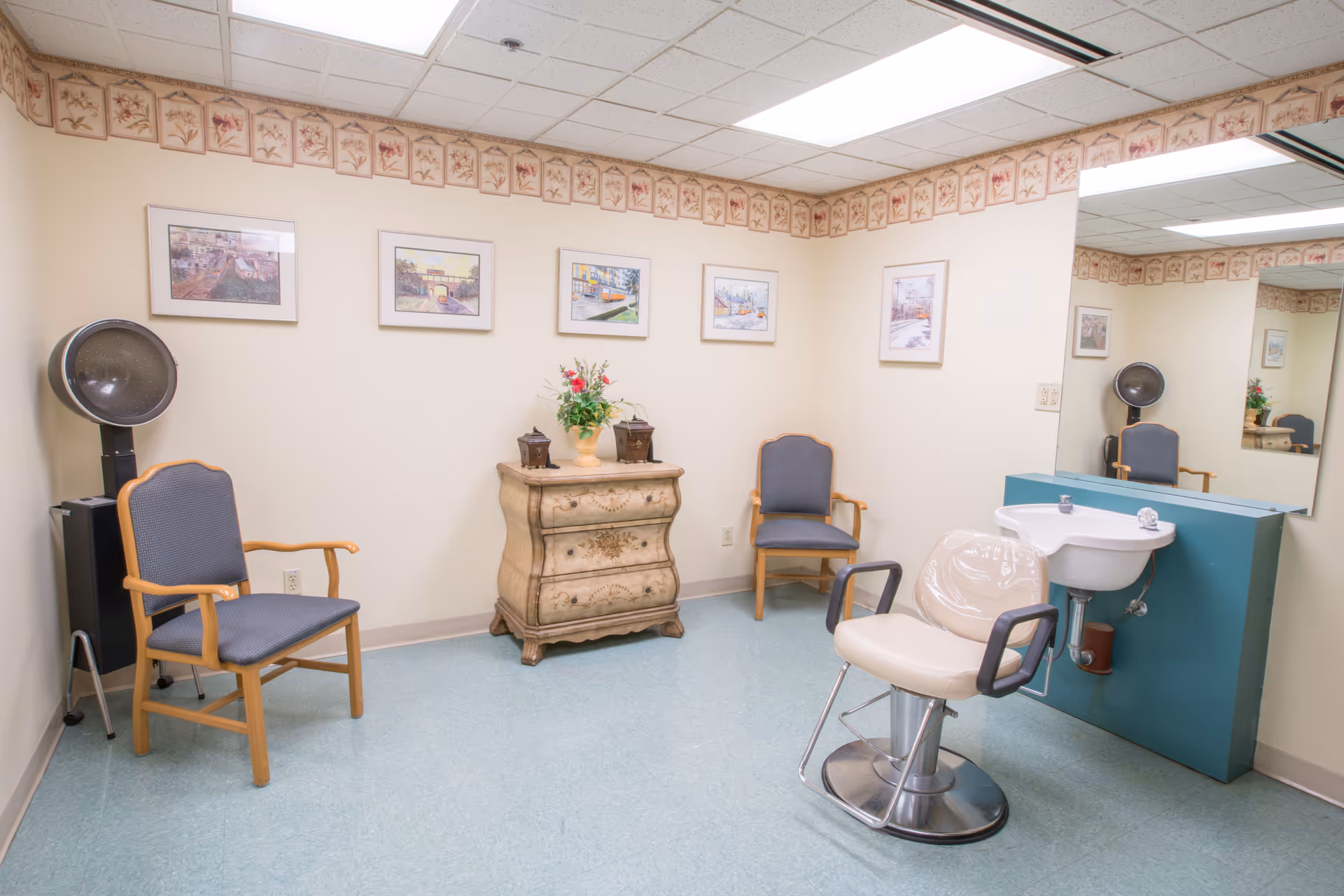 A small salon room with a salon chair in front of a sink and mirror, two wooden chairs with blue cushions, a decorative chest with a flower arrangement on top, and framed pictures on the walls. The room has a light blue floor and floral wallpaper border near the ceiling.