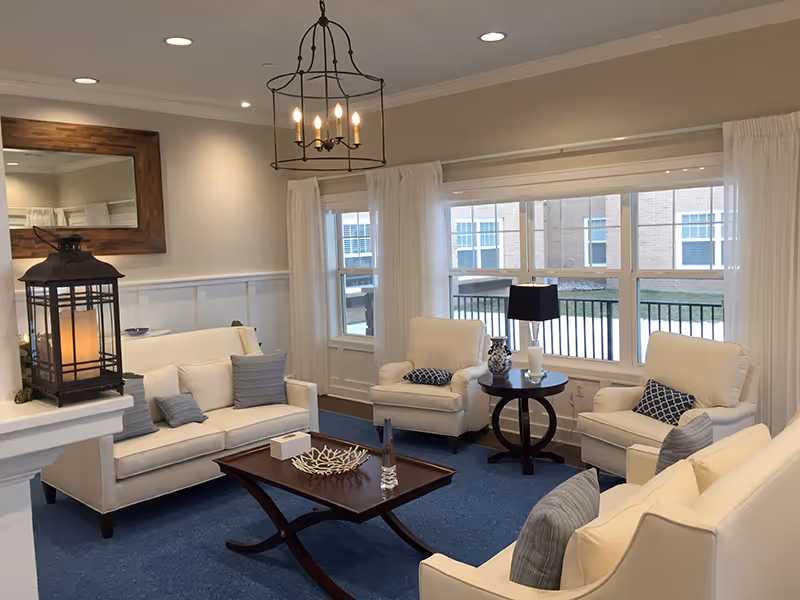 A bright and cozy living room with white upholstered sofas and armchairs arranged around a dark wooden coffee table. The room features large windows with white curtains, a blue carpet, a decorative lantern on a white mantel, a round side table with a lamp and decorative items, and a wooden-framed mirror on the wall. A chandelier with candle-style lights hangs from the ceiling.
