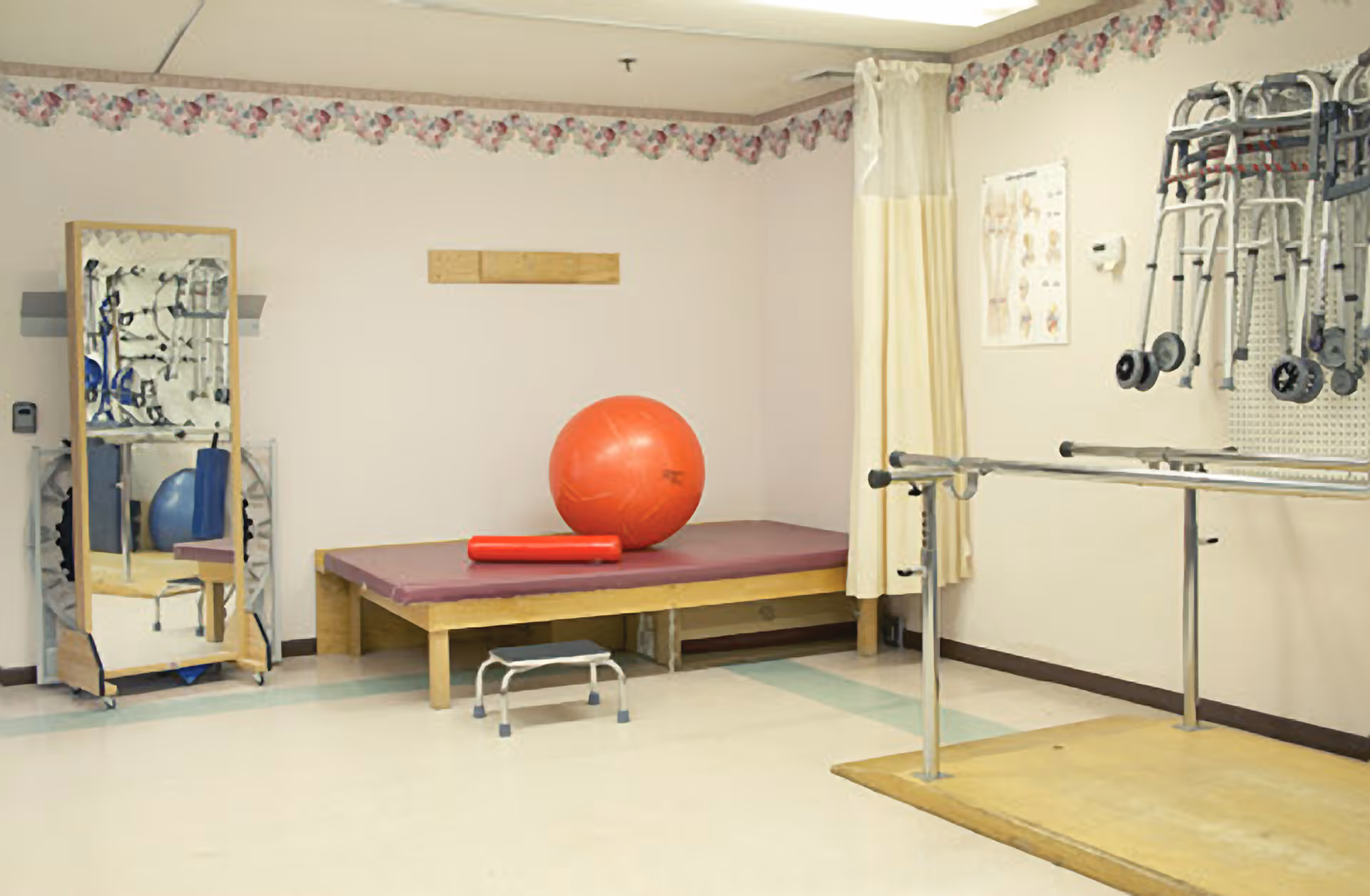 A physical therapy room with a padded treatment table holding a large red exercise ball and a red foam roller. There is a full-length mirror on a wooden stand, a small step stool on the floor, parallel bars for walking exercises, and several walkers hanging on the wall. The room has light-colored walls with a floral border near the ceiling and a curtain partially covering one corner.
