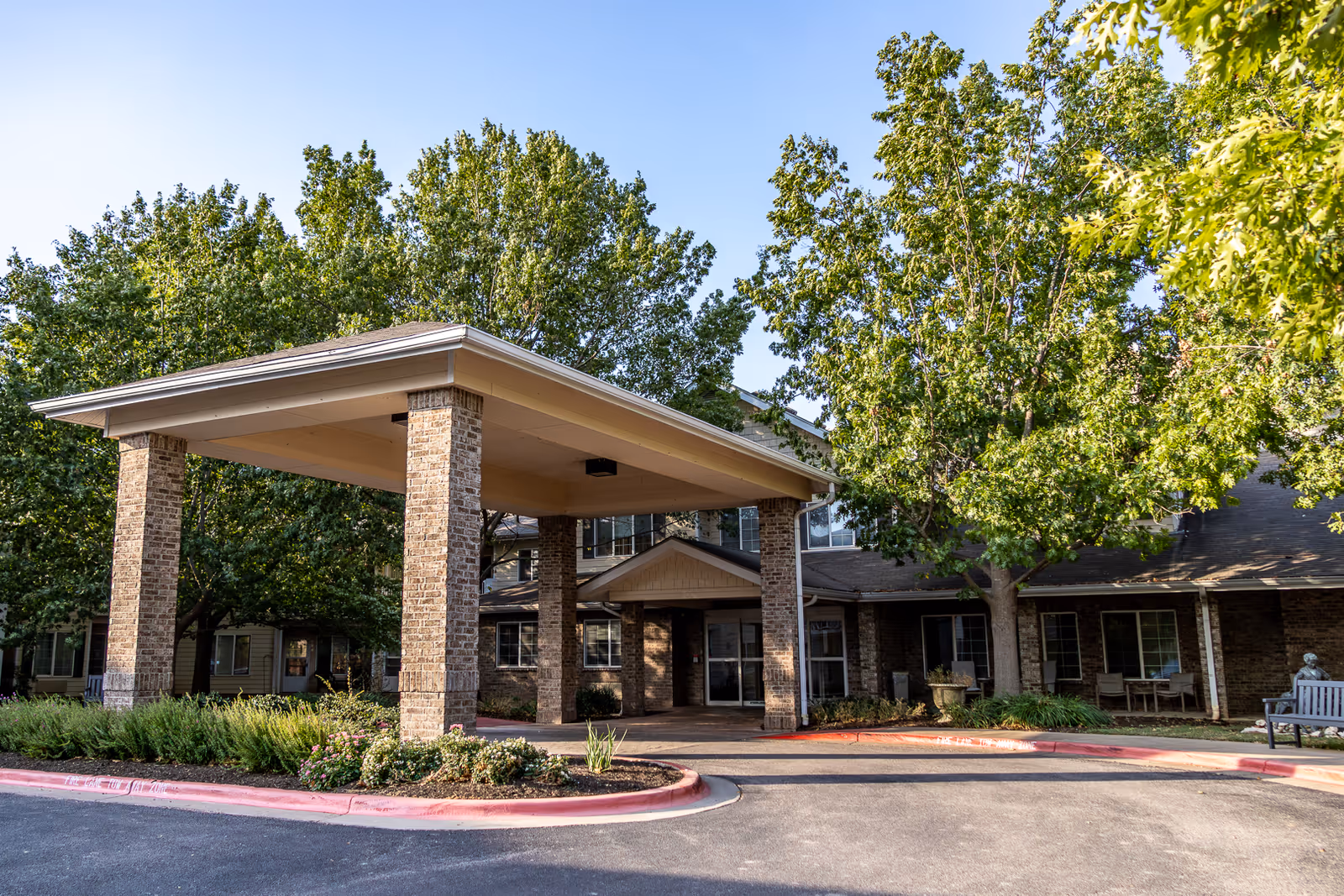 Covered porte-cochère entrance to a brick senior living building surrounded by trees and landscaping.
