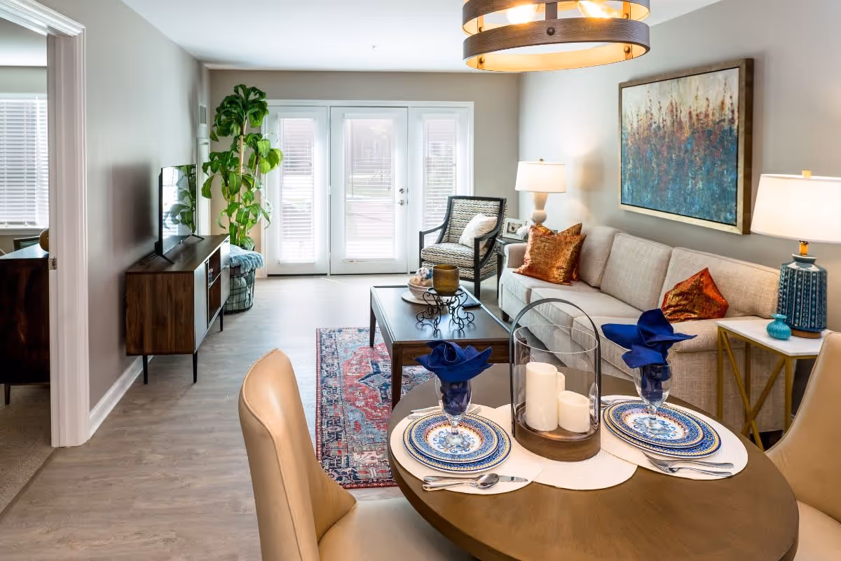 A cozy living room and dining area with a round wooden table set for two with blue napkins and decorative plates. The living room features a beige sofa with orange pillows, a patterned armchair, a wooden coffee table, a colorful rug, a large plant near glass doors with blinds, and two table lamps. A large abstract painting hangs on the wall above the sofa.