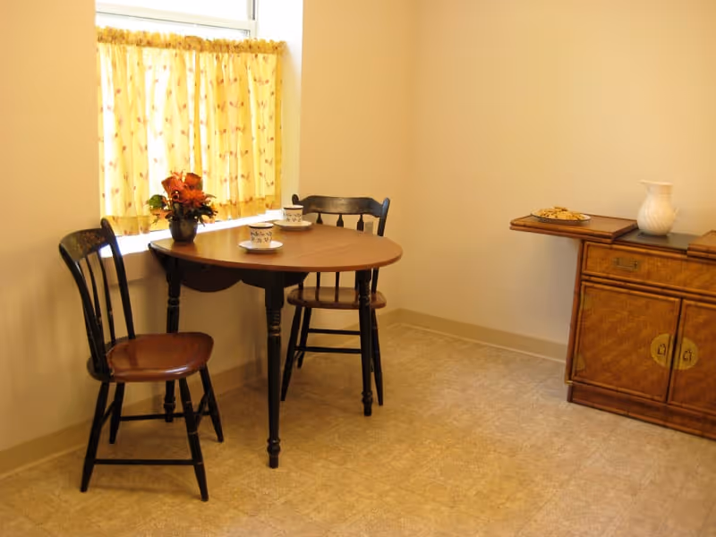 Small dining nook with a round wooden table, two chairs, a window with yellow curtains, and a side cabinet.