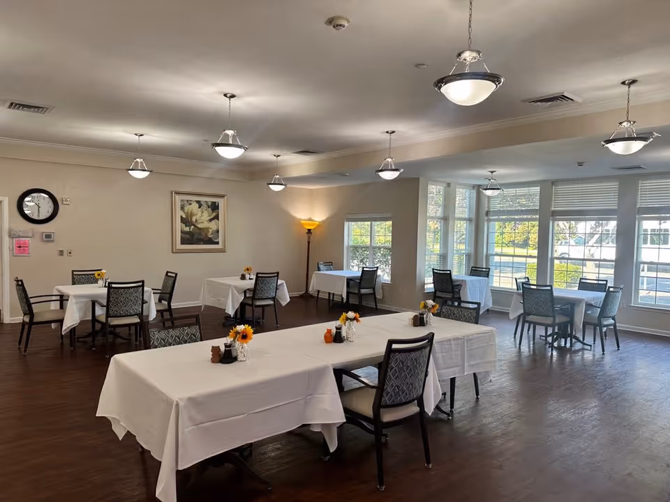 A bright dining room with several tables covered in white tablecloths, each set with chairs and small flower arrangements. Large windows let in natural light, and ceiling lights hang evenly spaced across the room. A clock and a framed floral painting are visible on the wall.