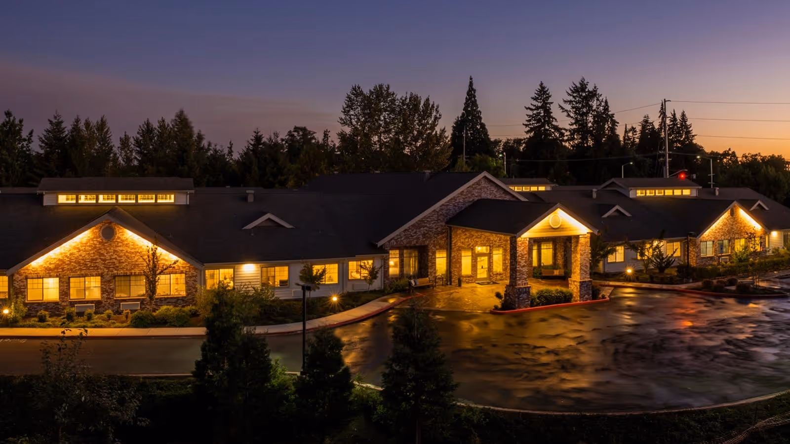 Exterior front of a single-story memory care facility lit up at dusk with a circular driveway and landscaped grounds.