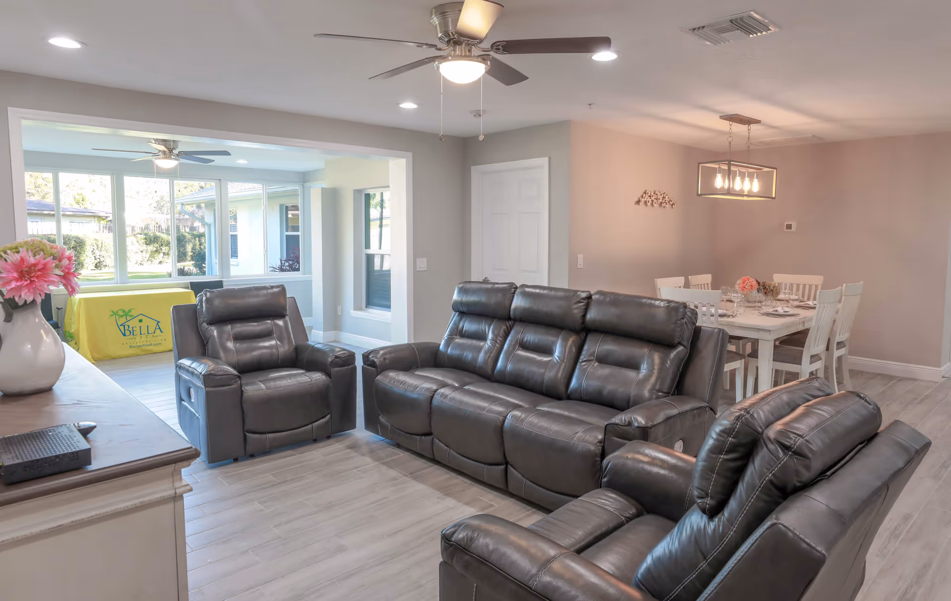 A bright and modern living room with gray leather recliner sofa and chairs, a ceiling fan with light, and a dining area with a white table and chairs in the background. Large windows and a door lead to a sunroom with more seating and a yellow tablecloth with the Bella Vita logo.