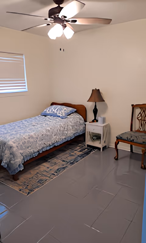 A simple bedroom with a single bed covered in a blue patterned bedspread and matching pillow. Next to the bed is a small white nightstand with a black lamp and a tissue box. There is a wooden chair with a cushioned seat against the wall. The floor is tiled and there is a ceiling fan with lights above. A window with a closed blind is on the left wall.