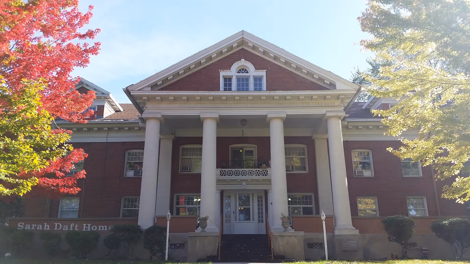 Front exterior view of a large, historic brick building with white columns and a triangular pediment. The building is surrounded by trees with autumn foliage, including vibrant red and yellow leaves. A sign near the entrance reads 'Sarah Daft Home.'