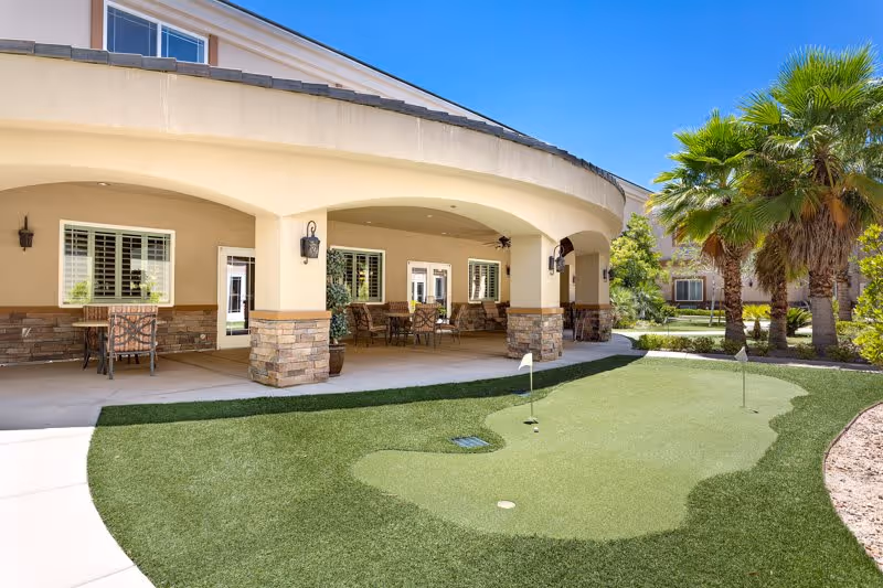 Outdoor patio area at San Martin Senior Living with a covered seating area featuring tables and chairs, adjacent to a small putting green with two golf holes and flags, surrounded by palm trees and landscaping.