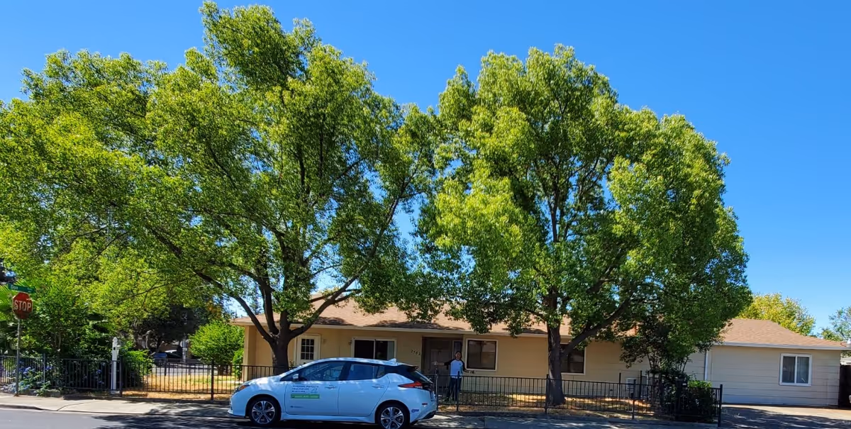 Single-story residential building with a beige exterior and brown roof, partially shaded by two large green trees in front. A white car is parked on the street in front of the building, and a person is standing near the entrance. The sky is clear and blue.