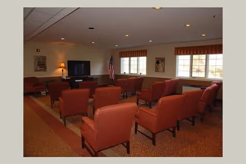 A common area with multiple red upholstered armchairs arranged in rows facing a television on a stand. The room has beige walls, a carpeted floor, two windows with striped valances, an American flag in the corner, and a lamp on a side table.