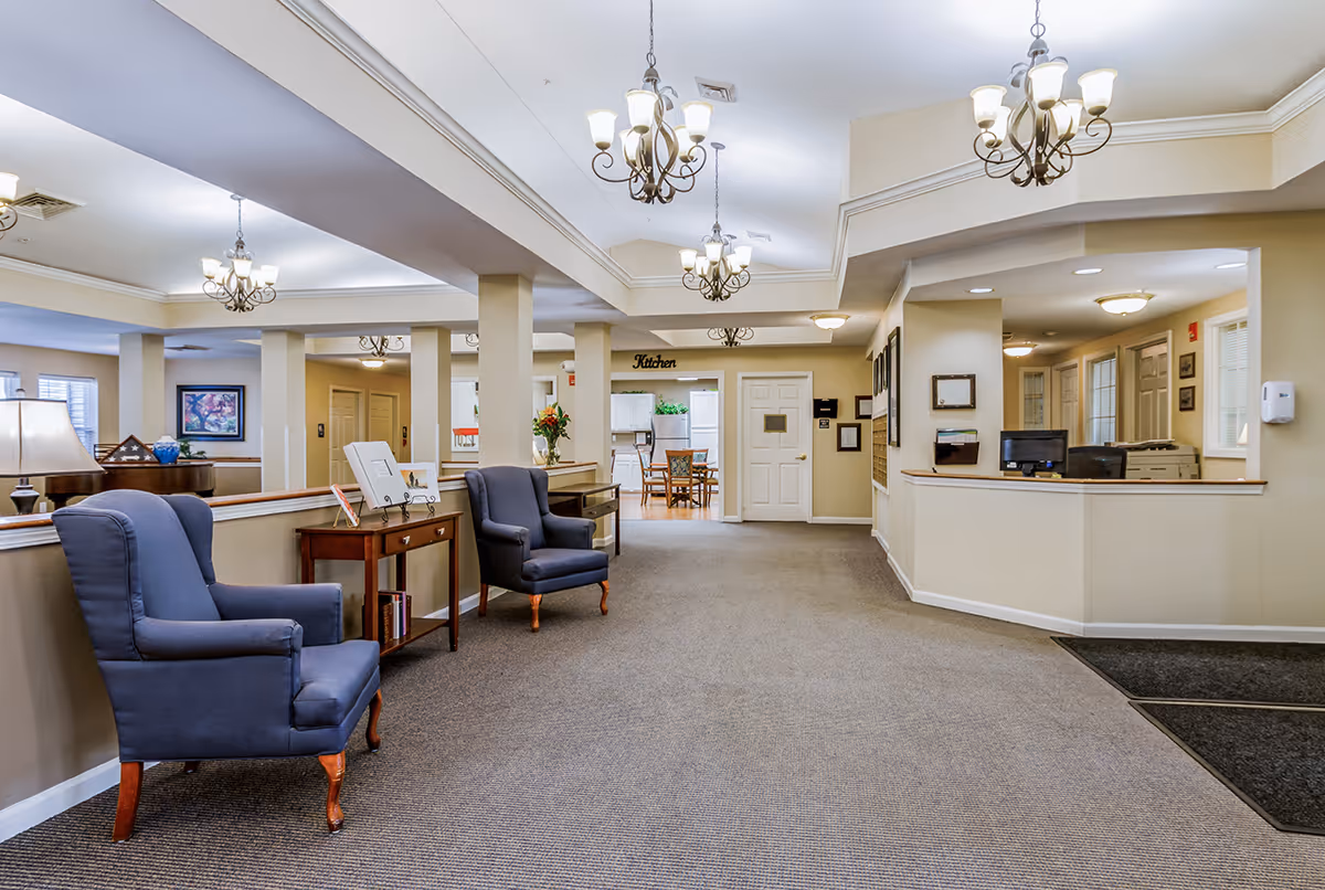 Interior view of a senior living facility lobby area with beige walls and carpeted floor. There are two blue upholstered armchairs and a small wooden table with books and decorative items. Multiple chandeliers hang from the ceiling. In the background, there is a reception desk with computer monitors and a doorway labeled 'Kitchen' leading to a dining area with a table and chairs.