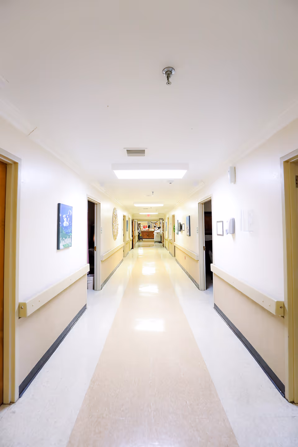 Well-lit long nursing home corridor with handrails, doors and artwork along both walls.