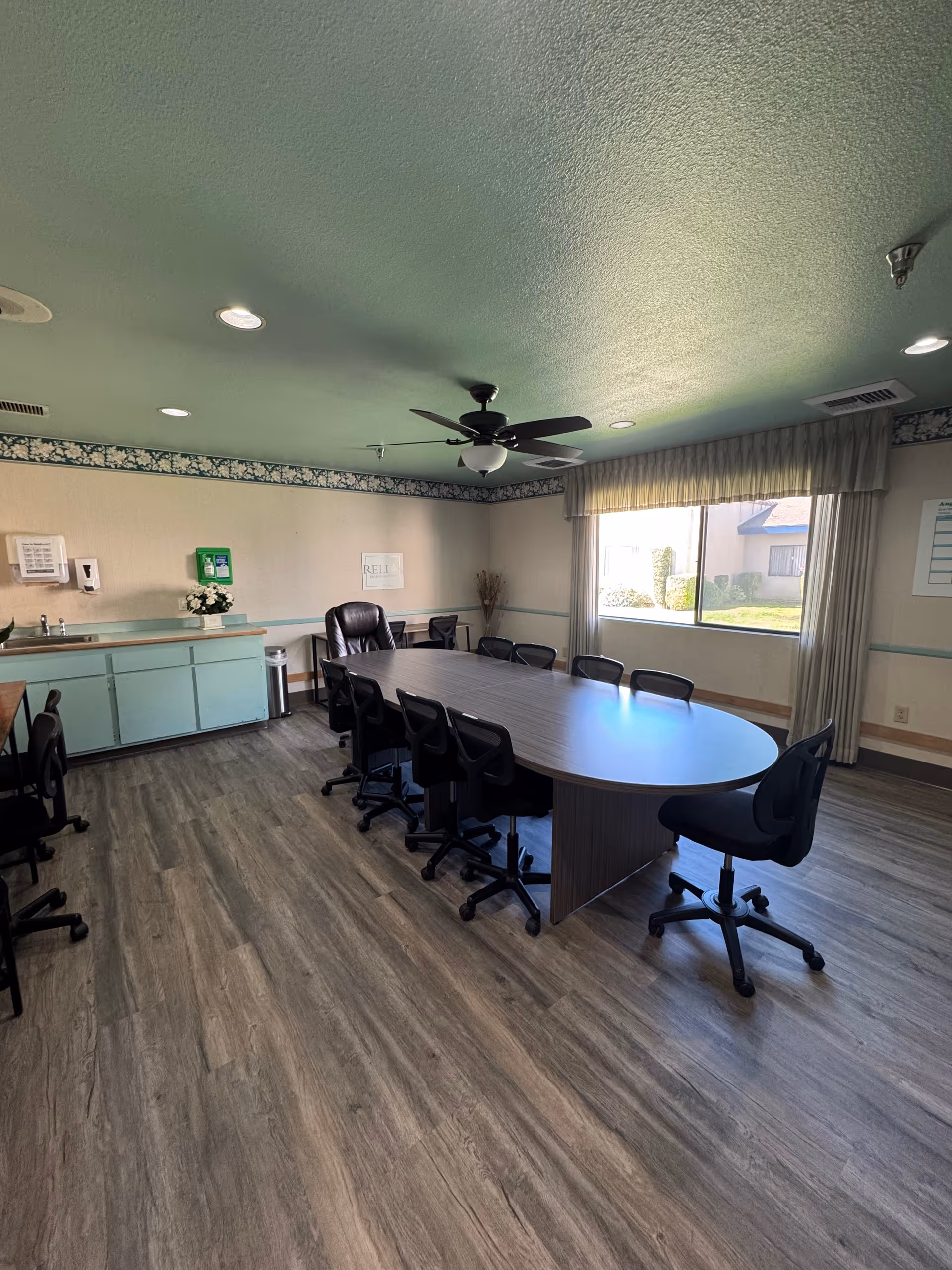 A meeting room with a long oval conference table surrounded by rolling office chairs, a ceiling fan, window, and a small kitchenette area.