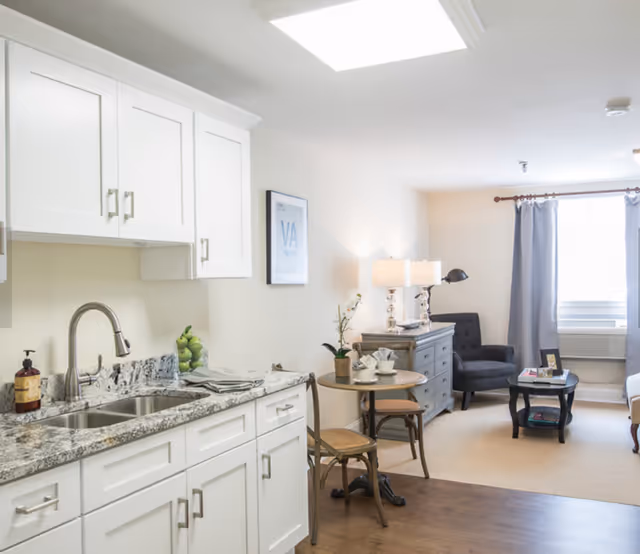 Bright open-plan apartment interior showing a small kitchen with white cabinets and a granite countertop opening to a cozy living area with an armchair, side table, and window.