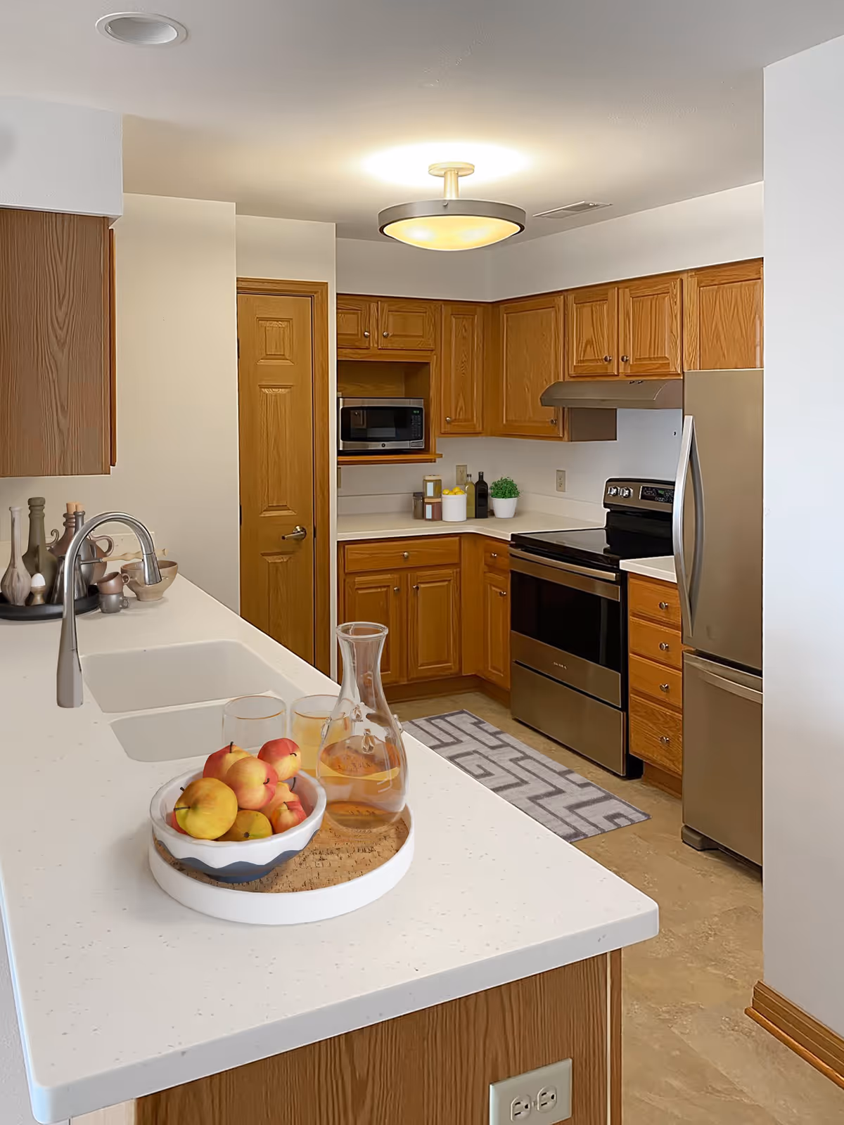 A modern kitchen with wooden cabinets, a stainless steel refrigerator and oven, a microwave, and a white countertop island with a sink. On the countertop, there is a tray holding a bowl of apples, a glass pitcher with a beverage, and two glasses. The kitchen has a ceiling light fixture and a patterned rug on the floor.