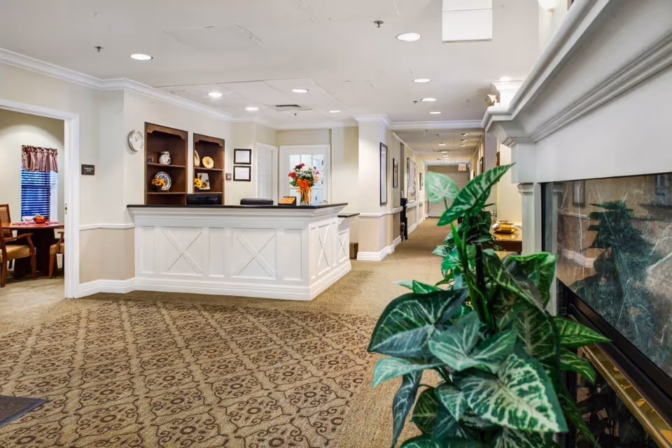 Reception area of a senior living facility with a white front desk, decorative shelves with vases and flowers, a patterned carpet, a hallway leading further into the building, and a fireplace with green plants in the foreground.