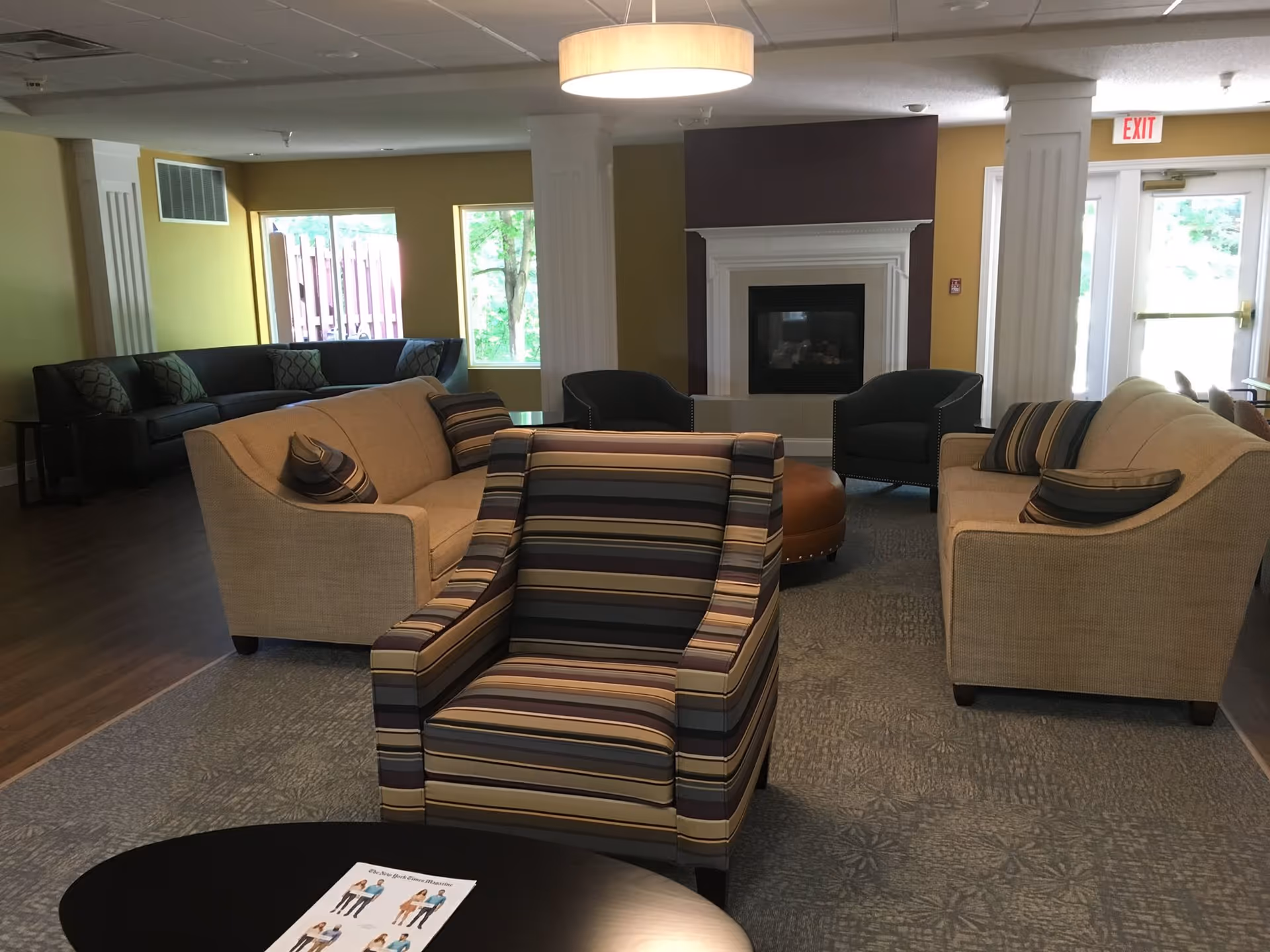 A cozy living room area in The Arbordale facility featuring multiple sofas and armchairs arranged around a central fireplace. The room has large windows letting in natural light, a round ottoman, and a modern ceiling light fixture. A table in the foreground holds a brochure.