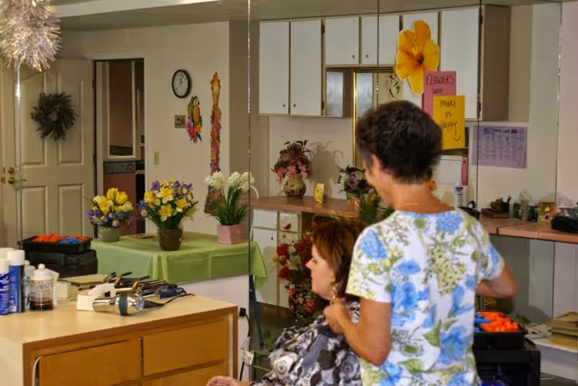 A woman standing behind another woman seated in a chair, possibly styling her hair, in a room with a counter and cabinets. The room has various flower arrangements on tables and counters, a clock on the wall, and colorful decorations including a sign that says 'FLOWERS will MAKE US HAPPY'.