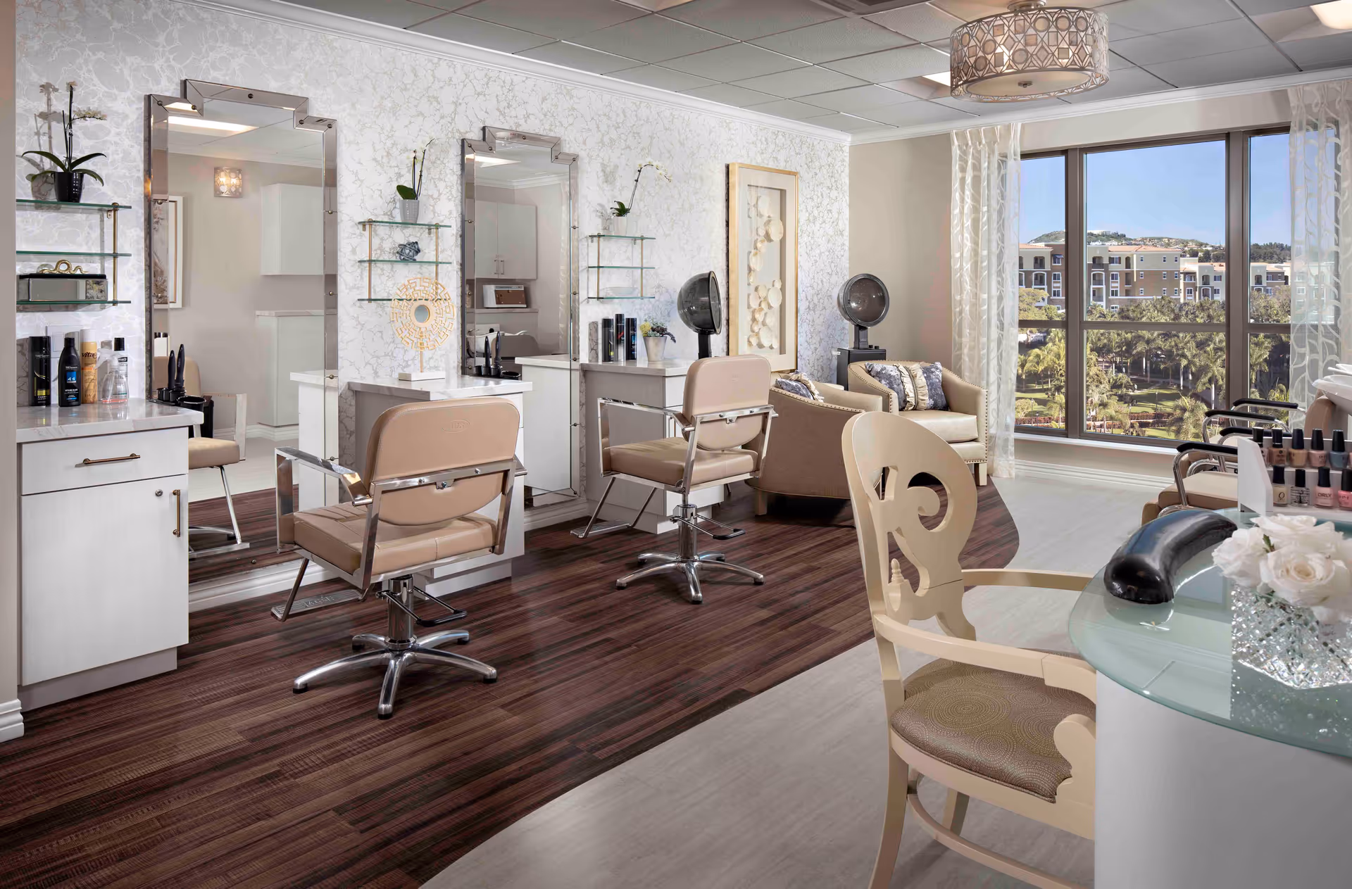 A bright and elegant salon area in a senior living facility featuring two styling chairs in front of large mirrors, a manicure table with nail polish and a UV nail dryer, a cozy seating area with armchairs and a large window showing an outdoor view of buildings and greenery.