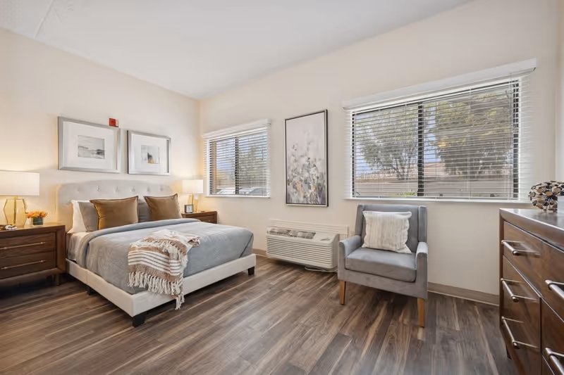 A bright and cozy bedroom with a large bed featuring a tufted headboard, gray bedding, and brown accent pillows. Two wooden nightstands with lamps flank the bed. The room has two windows with blinds, a gray armchair with a cushion, a wooden dresser, and framed artwork on the walls. The floor is wood with a warm tone.