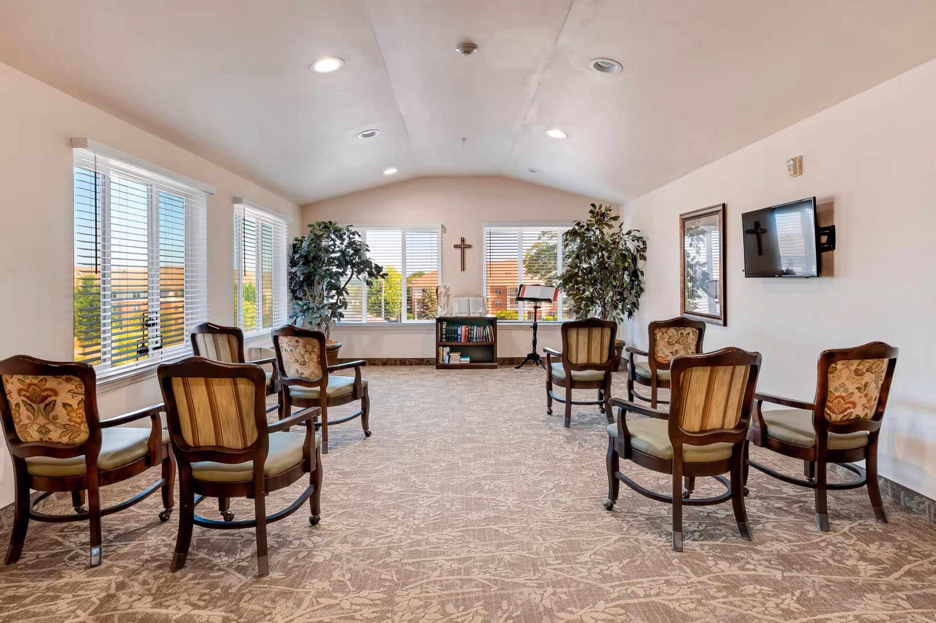 A bright room with large windows and beige walls, featuring eight wooden chairs with cushioned seats arranged in a circle on a patterned carpet. There are two large potted plants near the windows, a small bookshelf with books, a music stand, a wall-mounted TV, a framed picture, and a cross on the wall.