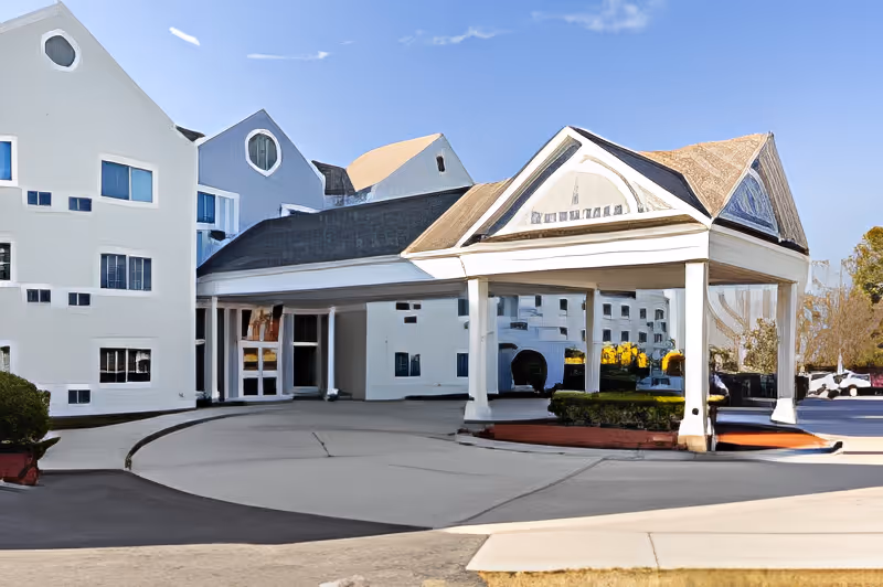 Exterior view of a senior living facility with a covered entrance supported by white columns, a driveway for vehicles, and a multi-story building with multiple windows under a clear blue sky.