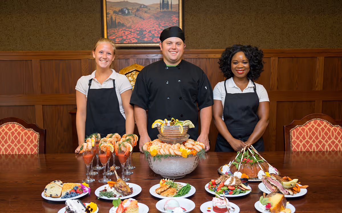 Three smiling dining staff (two servers and a chef) stand behind a long table of seafood and plated dishes in a formal dining room.