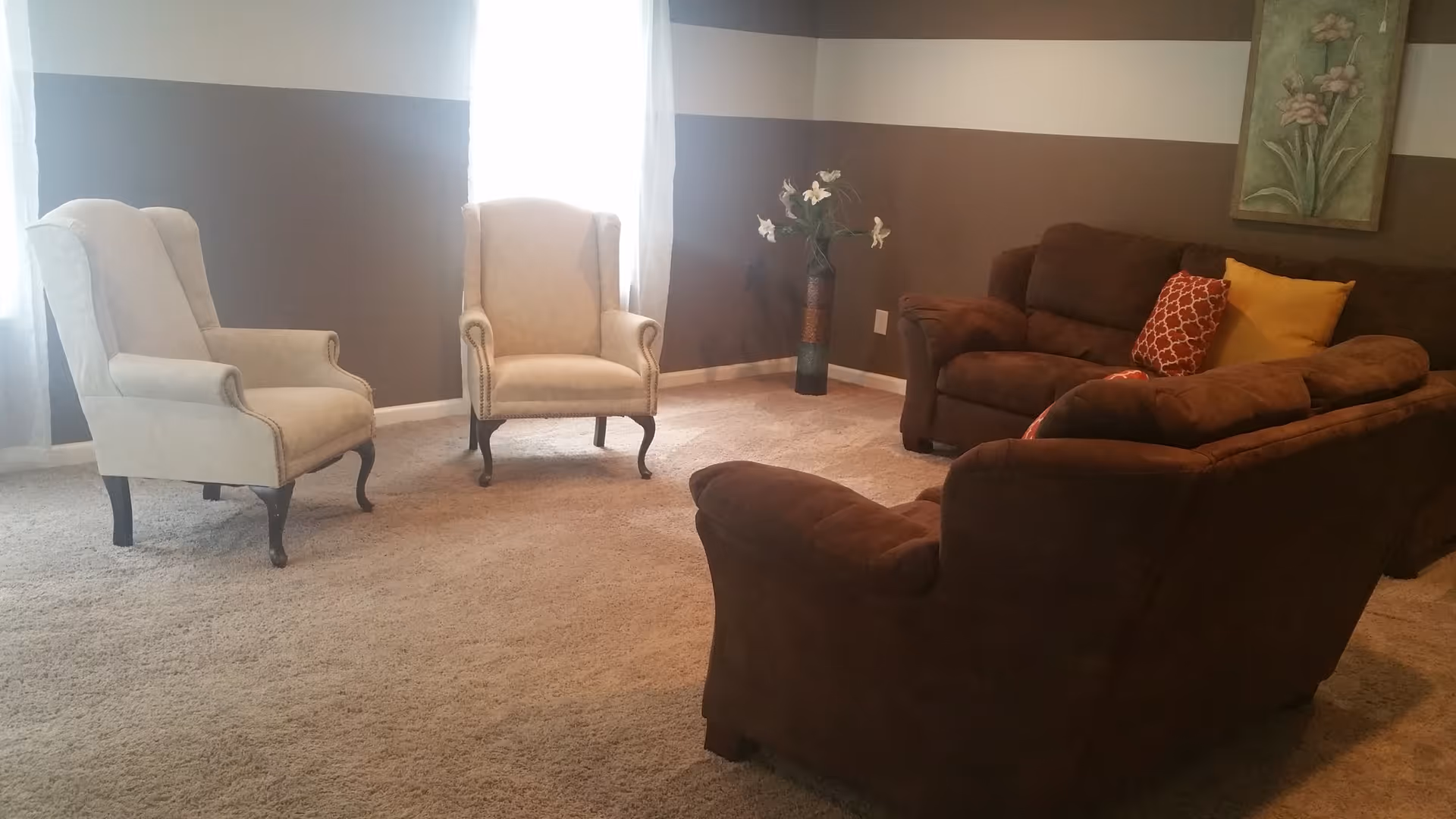 A cozy living room with two beige armchairs near two windows with white curtains, a brown sofa and loveseat with colorful throw pillows, a vase with flowers in the corner, and a floral painting on the wall.