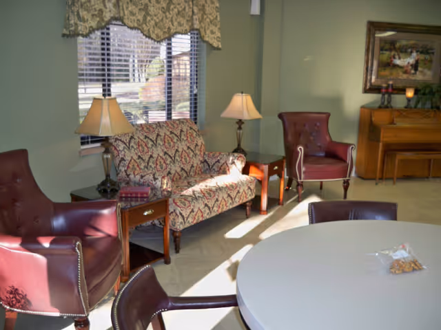 A cozy sitting area with two burgundy leather armchairs, a patterned loveseat, two wooden side tables with lamps, a round white table with chairs, and a piano against a light green wall with a framed painting above it. A window with blinds and a valance lets in natural light.
