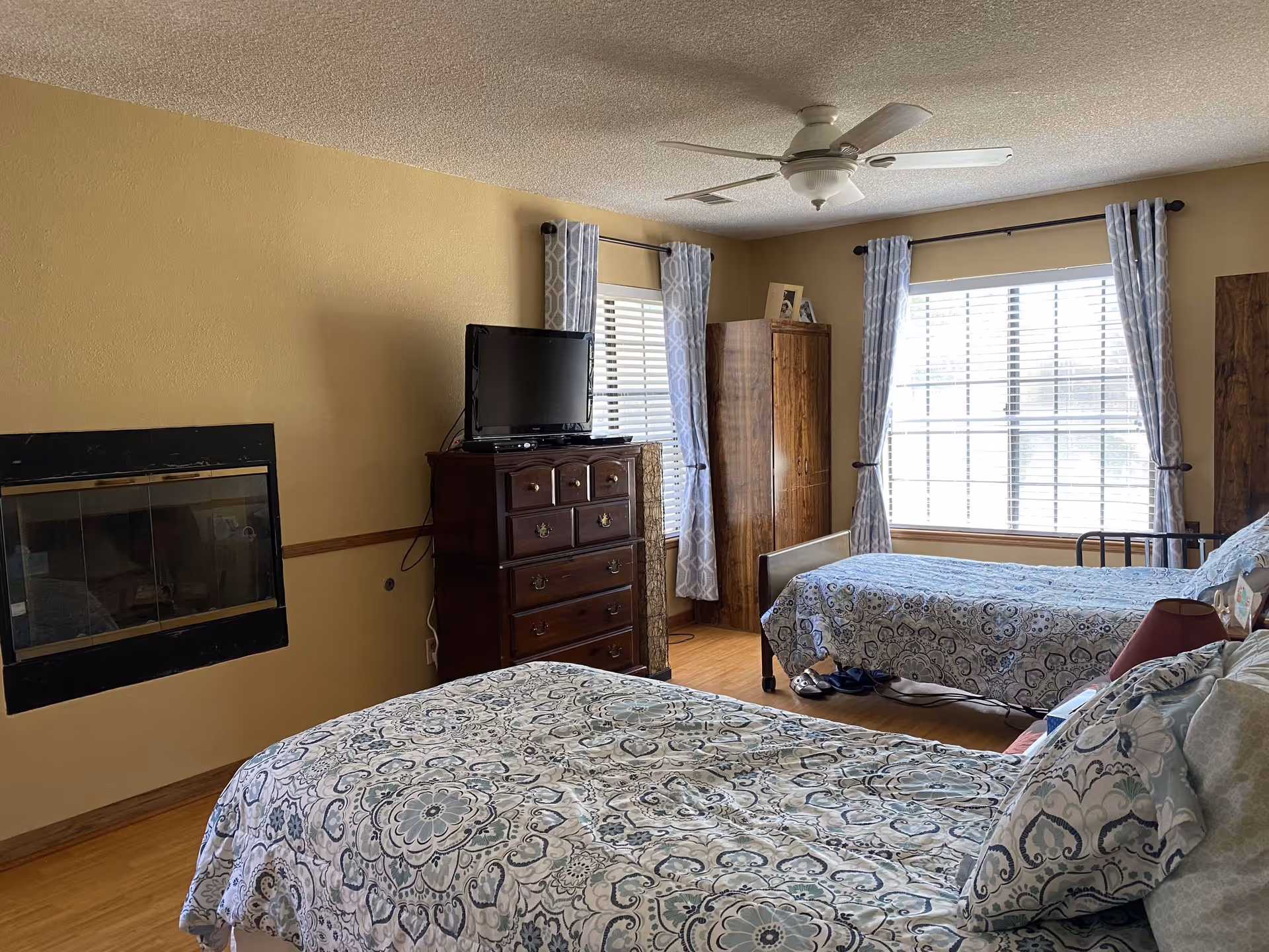 Bright shared bedroom with two twin beds, patterned bedding, a dresser with a TV, large windows with curtains, and a ceiling fan.