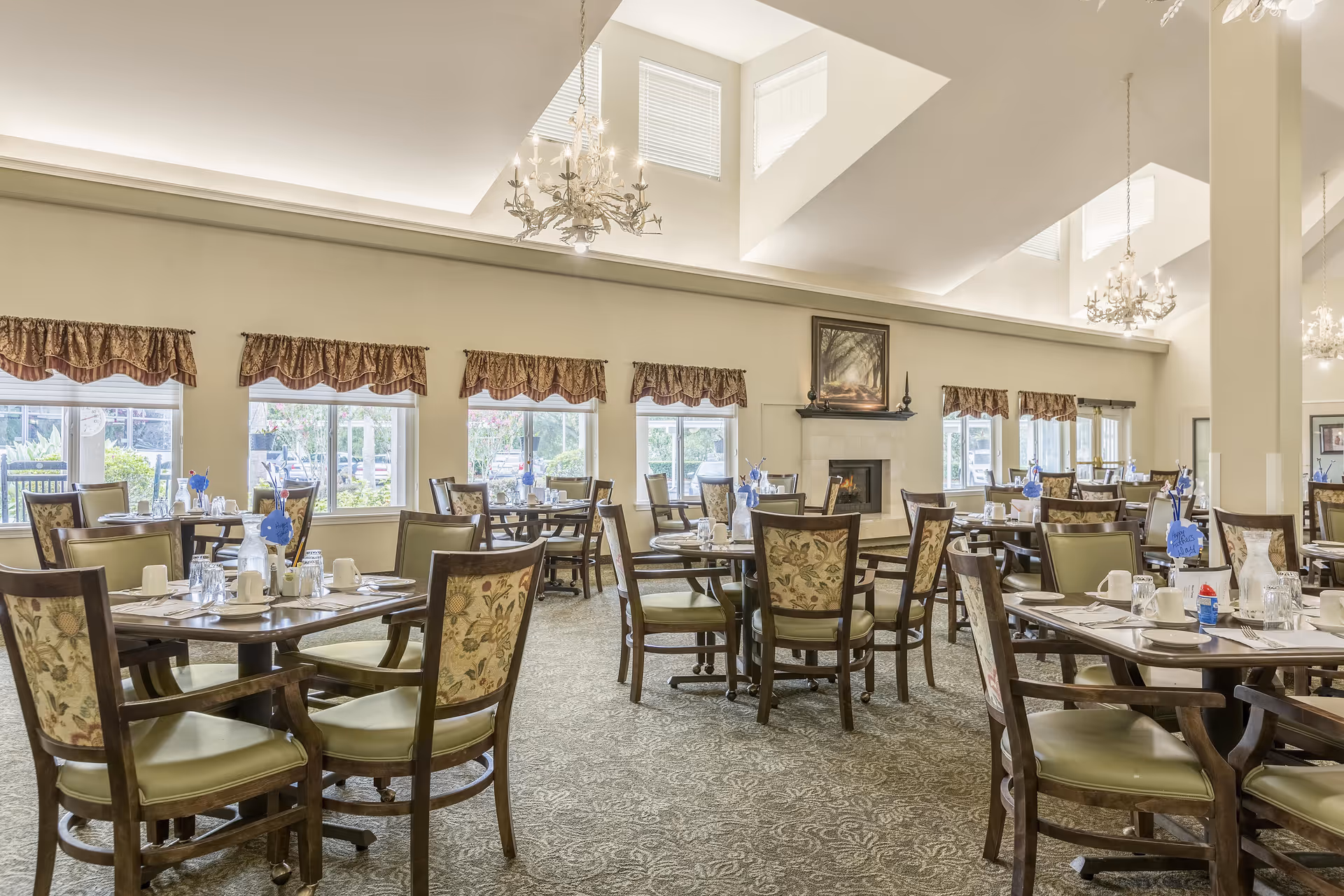 A bright and spacious dining room with multiple wooden tables and chairs arranged neatly. Each table is set with white cups, plates, and glasses, and decorated with small blue flower centerpieces. The room features large windows with patterned valances, a carpeted floor, and a fireplace with a framed painting above it. Elegant chandeliers hang from the high ceiling with skylights.