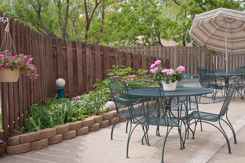 Outdoor patio with metal tables and chairs, a striped umbrella, potted pink flowers, and a wooden fence lining a planted garden bed.
