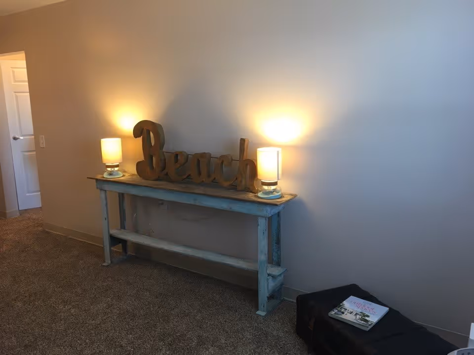 A cozy interior corner with a rustic blue wooden console table against a beige wall. On the table are two lit lamps with white shades and a decorative wooden sign spelling 'Beach'. To the right on the floor is a black ottoman with a book placed on top. A white door is visible in the background on the left.