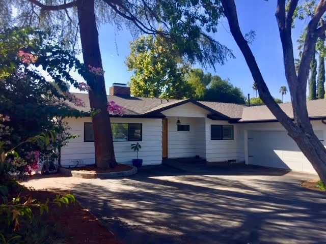 Single-story white ranch-style house with an attached garage, driveway, and trees casting shadows in front.