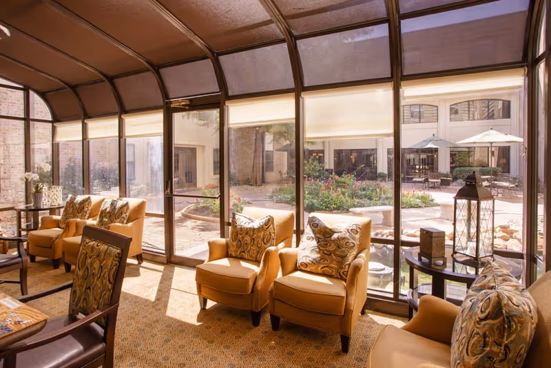 A sunlit indoor seating area with large curved glass windows overlooking a garden courtyard. The room features several cushioned armchairs with patterned pillows, a small round table with decorative items, and a carpeted floor. Outside, there are patio tables with umbrellas and a landscaped garden with flowers and greenery.