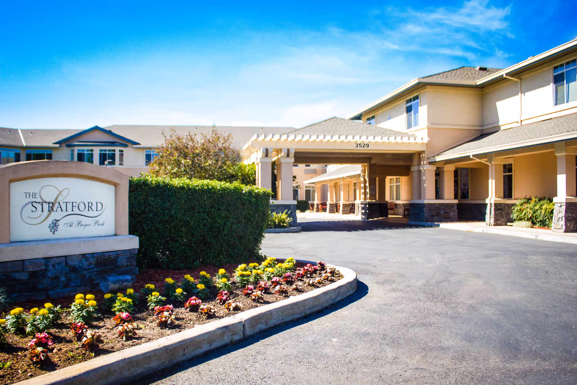 Exterior view of Stratford Beyer Park senior living facility with a driveway, landscaped flower beds, and a covered entrance. The building is beige with stone accents and multiple windows under a clear blue sky.